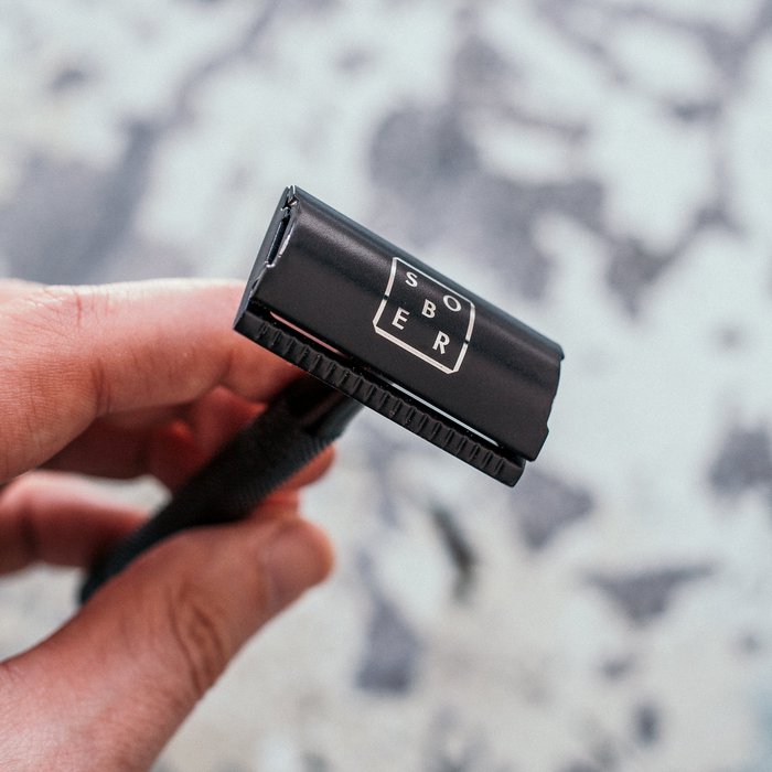 Hand houdt een zwarte safety razor met het merklogo SOBER in witte letters tijdens het scheren