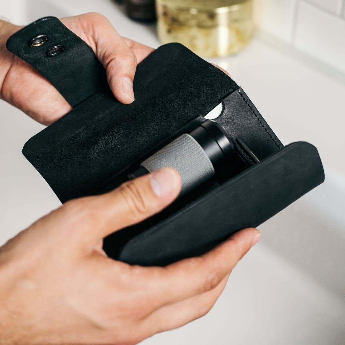 Handen die een zwarte leren etui openen met een compact apparaat erin, tegen een lichte achtergrond