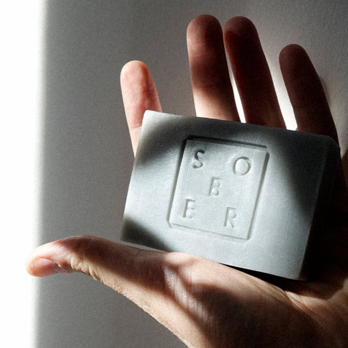 Hand met een grijze blokzeep met Sober logo in natuurlijk licht op grijze achtergrond