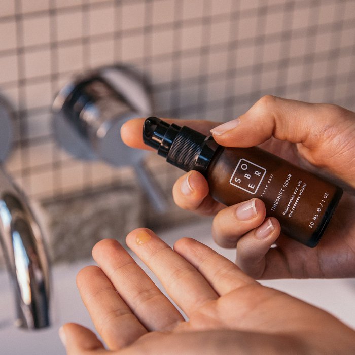 Hand die Sober Timeshift Serum doseert in badkamersetting met tegelachtergrond