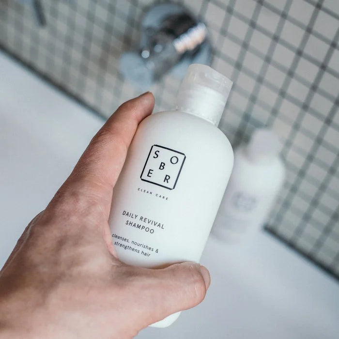 Hand houdt Sober Daily Revival Shampoo flesje vast in moderne badkamer met tegelwand