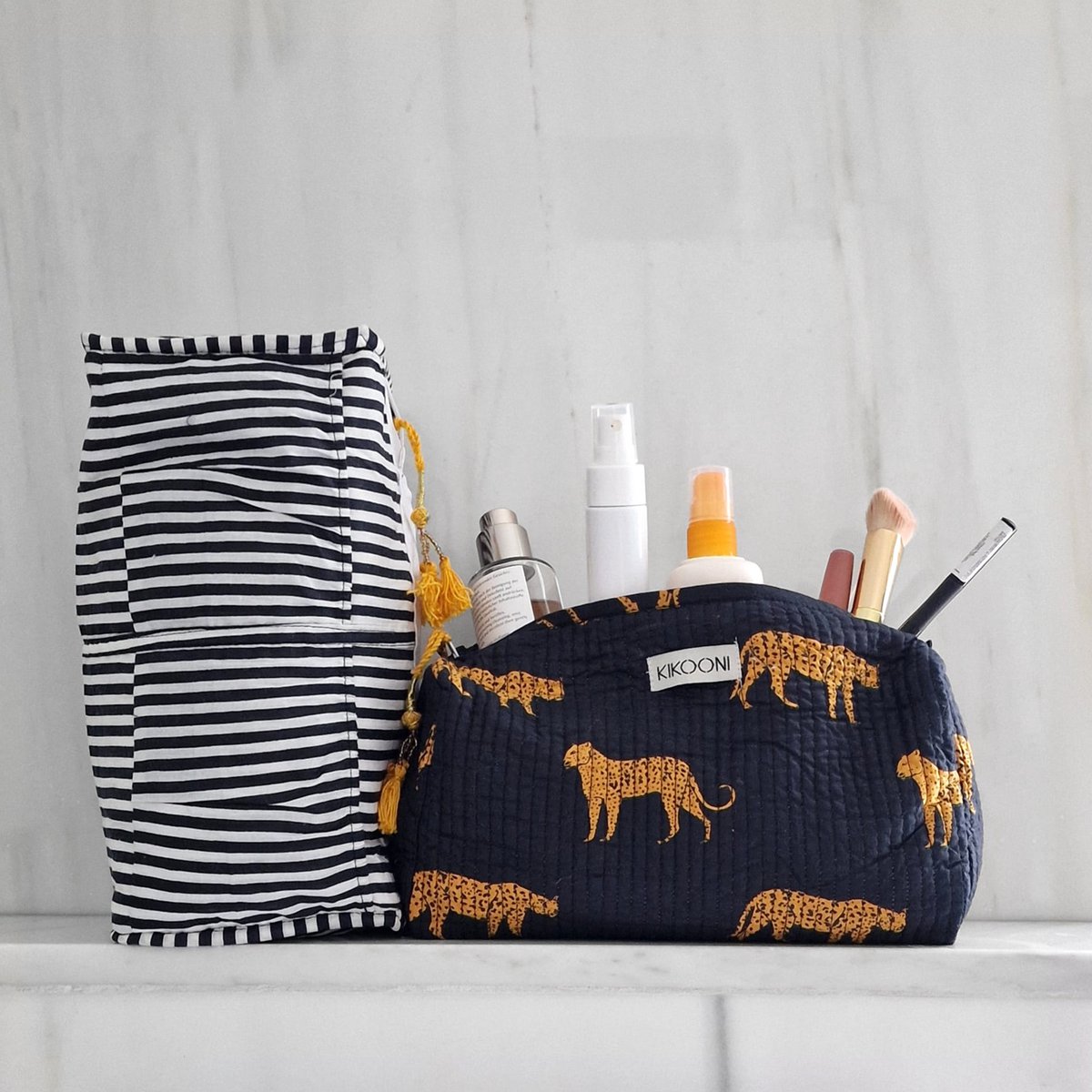 Zwart cosmetics tas met gele dierenprint en make-up producten naast gestreepte toilettas op marmeren ondergrond