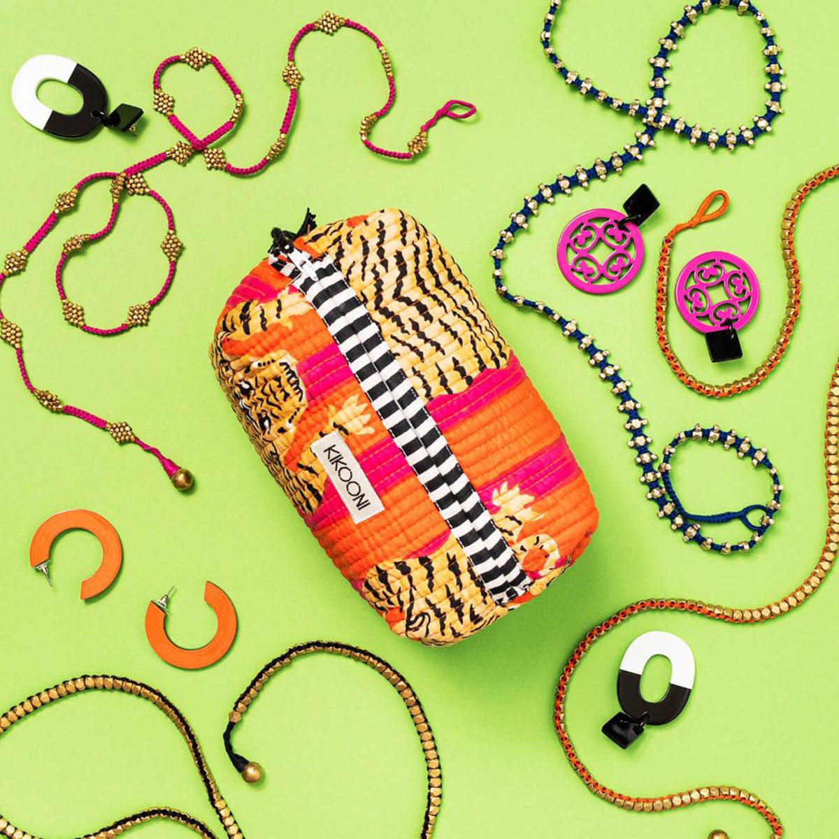 Felgekleurde sieraden en oorbellen rondom een oranje en roze patroon tasje met het label Kikcooni op groene achtergrond