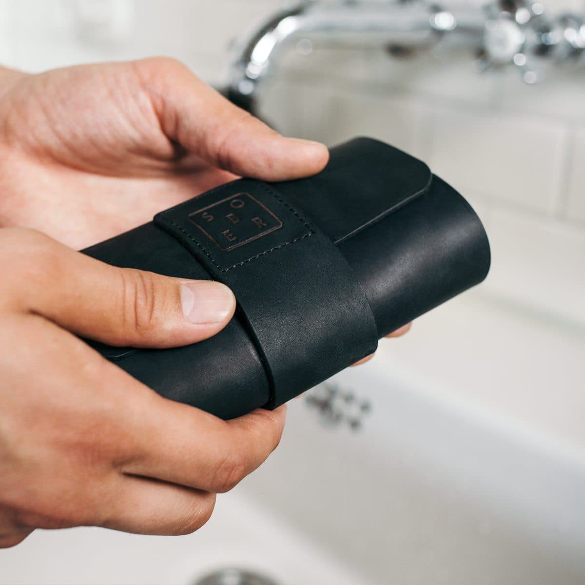 Hand houdt zwarte leren etui met SOER logo bij een witte wastafel en kraan