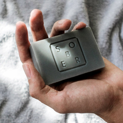 Hand houdt zwarte zeepblok met tekst 'S O B E R' op grijze achtergrond