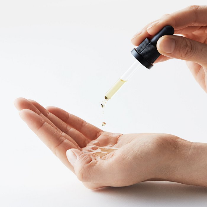 Hand druppelt cosmetische olie of serum uit pipet op handpalm tegen witte achtergrond