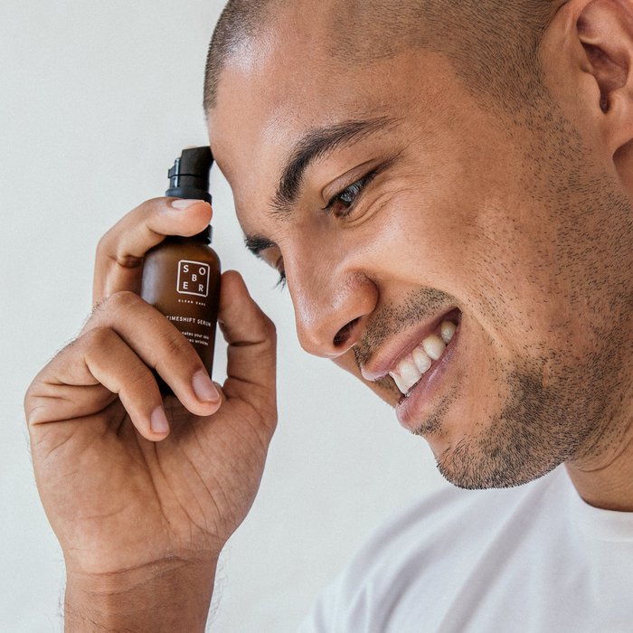 Man met kort haar en lichte baard houdt lachend Sober Timeshift serum gezichtsflesje vast