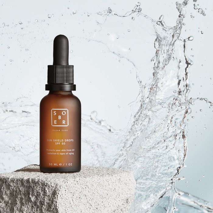 SOBER Sun Shield Drops SPF 50 in een bruine fles met pipet, water splash achtergrond